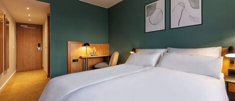 A hotel room with a large bed, a desk with a lamp, and two framed artworks on the wall.