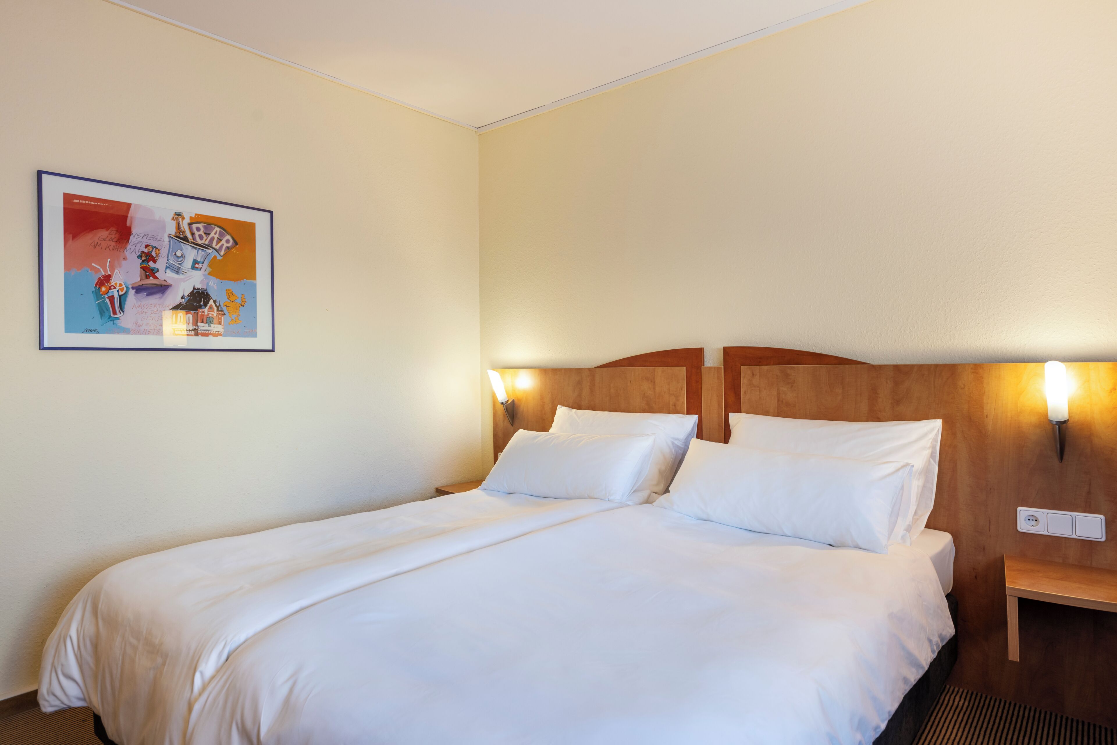 Une chambre d’hôtel avec un grand lit, deux tables de chevet, une lampe fixée au mur et un tableau encadré.