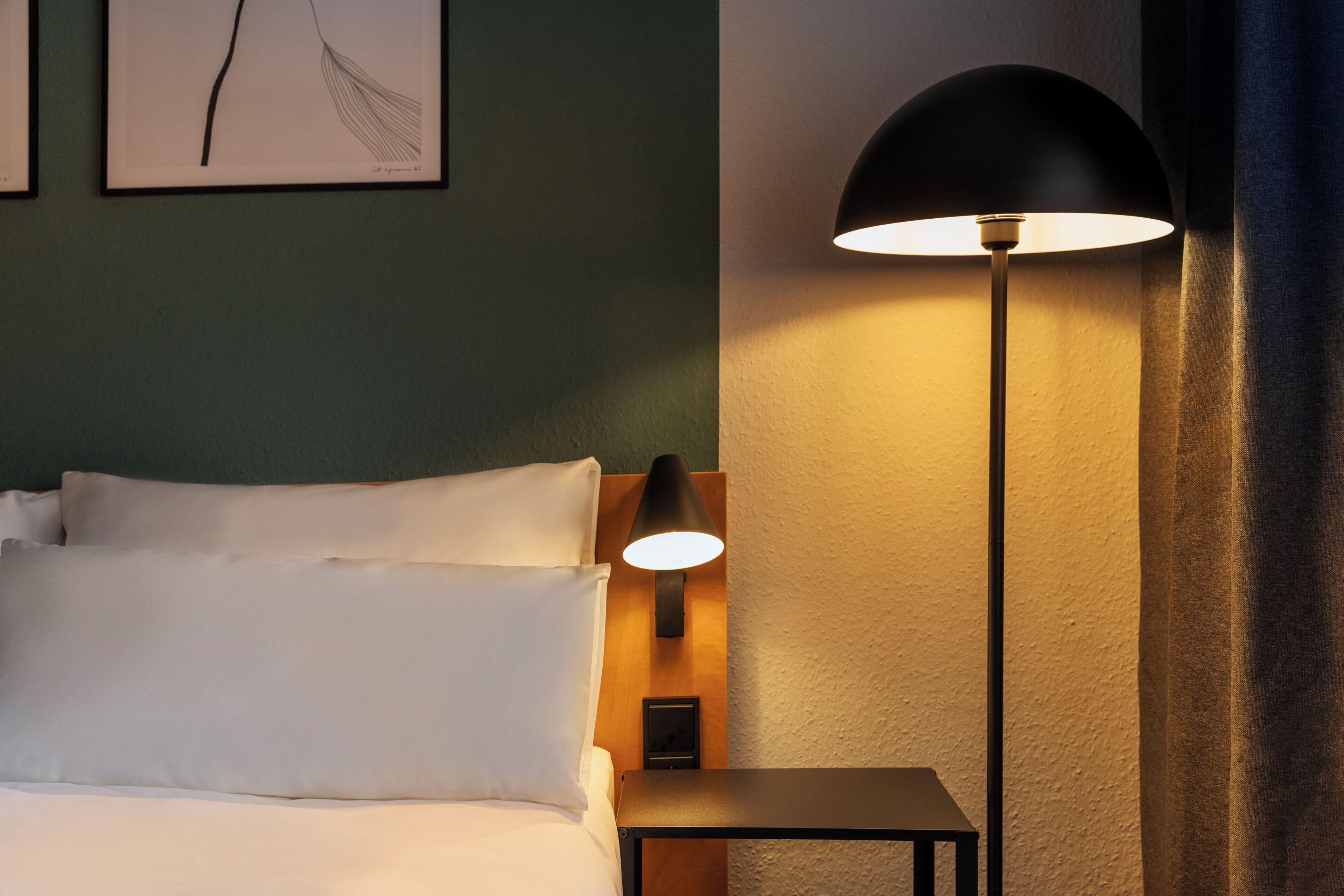 A neatly made bed with white linens, a black floor lamp, and a framed picture on the wall.