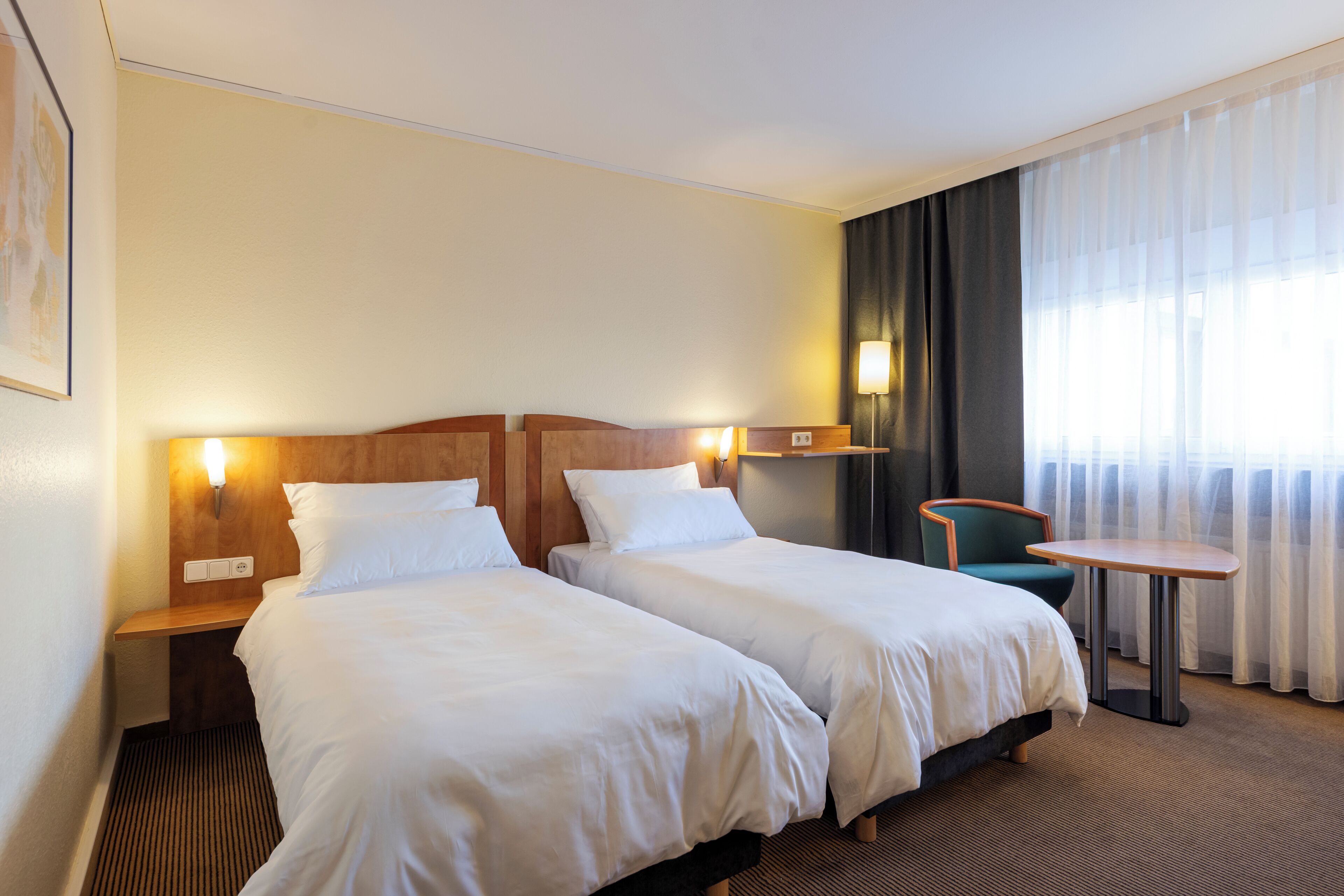 A hotel room with two beds, a small table, a chair, and a window with curtains.