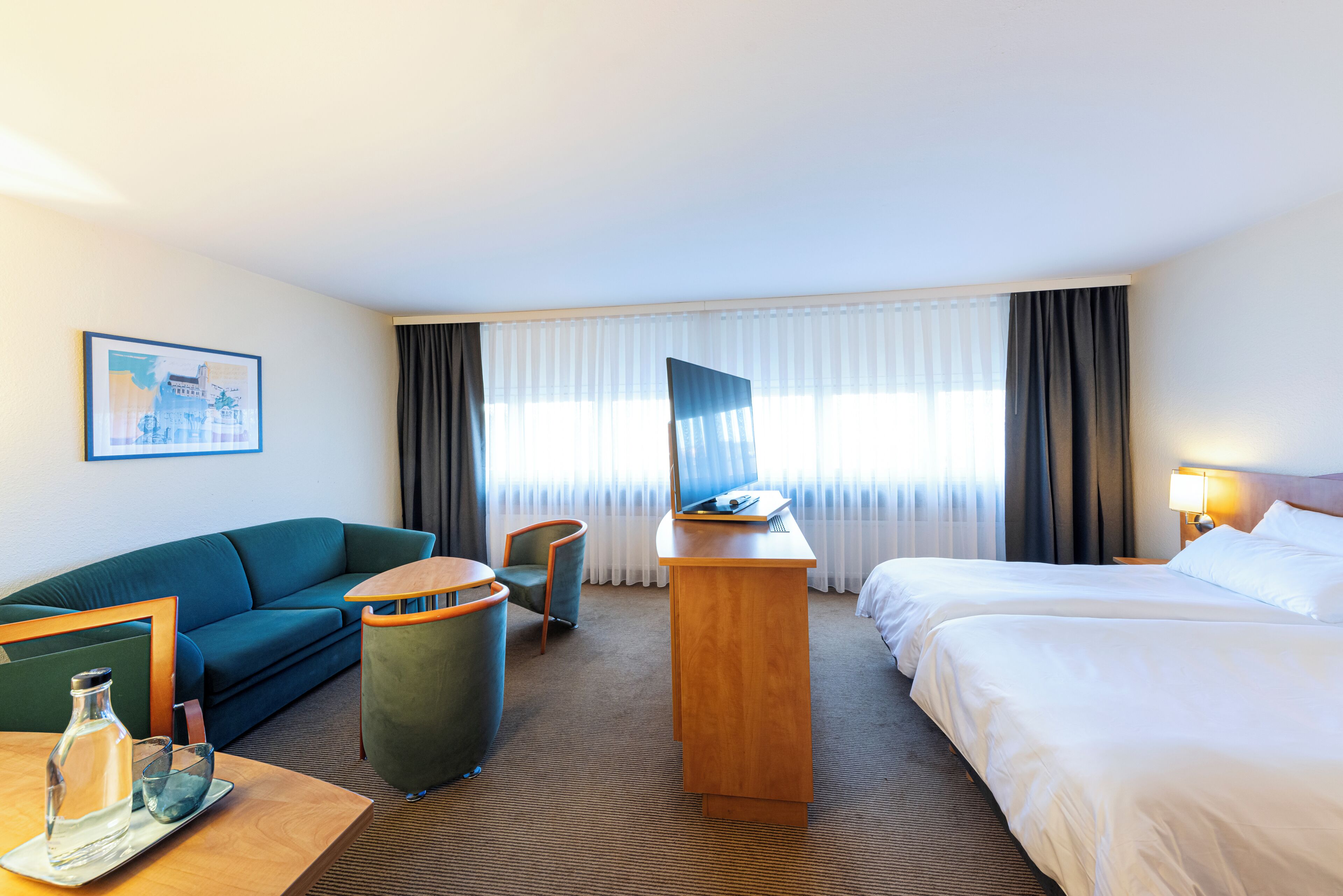 A hotel room with a bed, a desk, a chair, a sofa, and a painting on the wall.