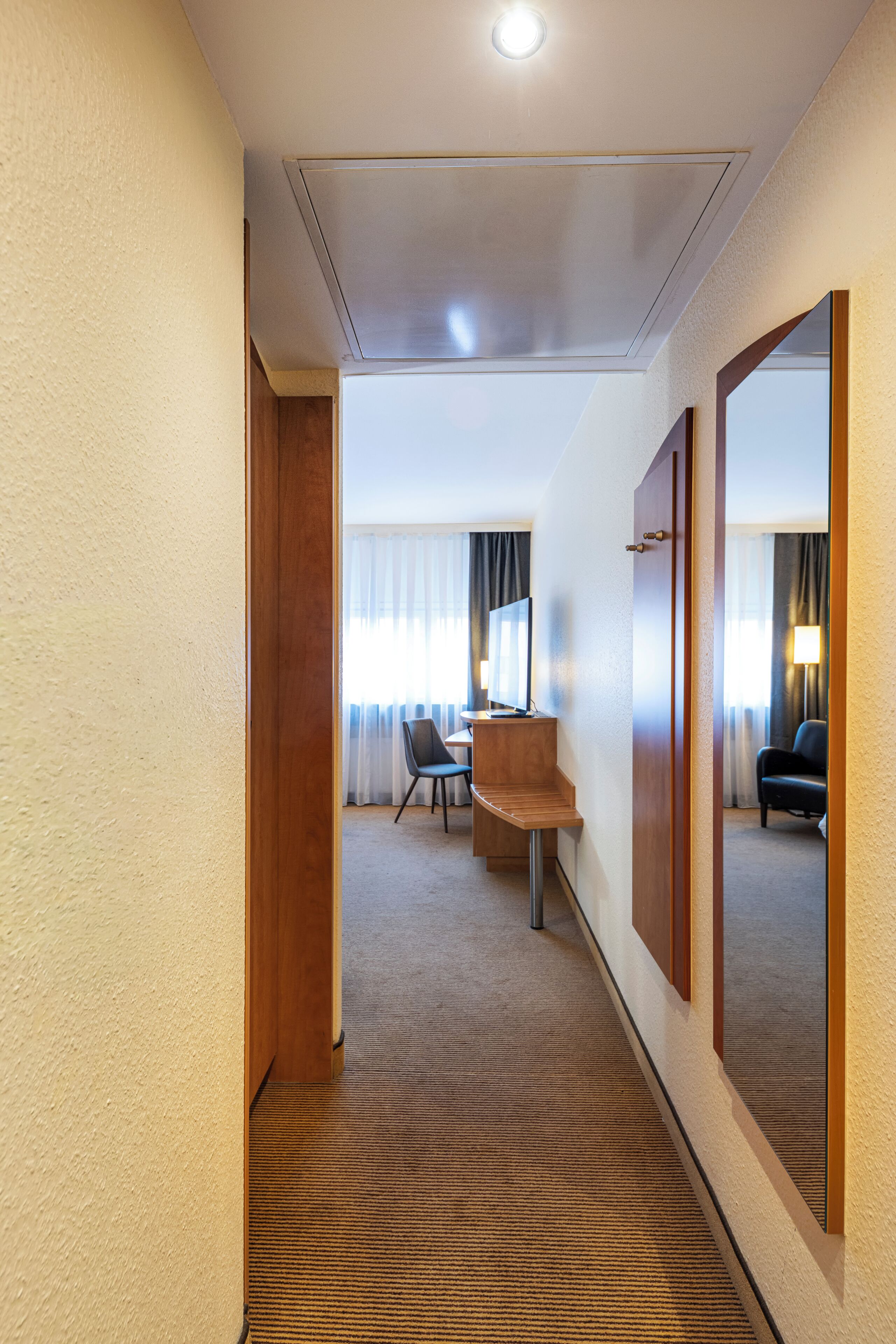 A hotel hallway with a carpeted floor, a wooden door, a desk with a chair, a computer, and a large mirror.