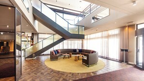 Un hall d’entrée spacieux, agrémenté d’un grand canapé d’angle aux lignes courbes, d’un parquet à motifs en chevrons, d’un escalier en verre et d’une grande fenêtre avec des rideaux.