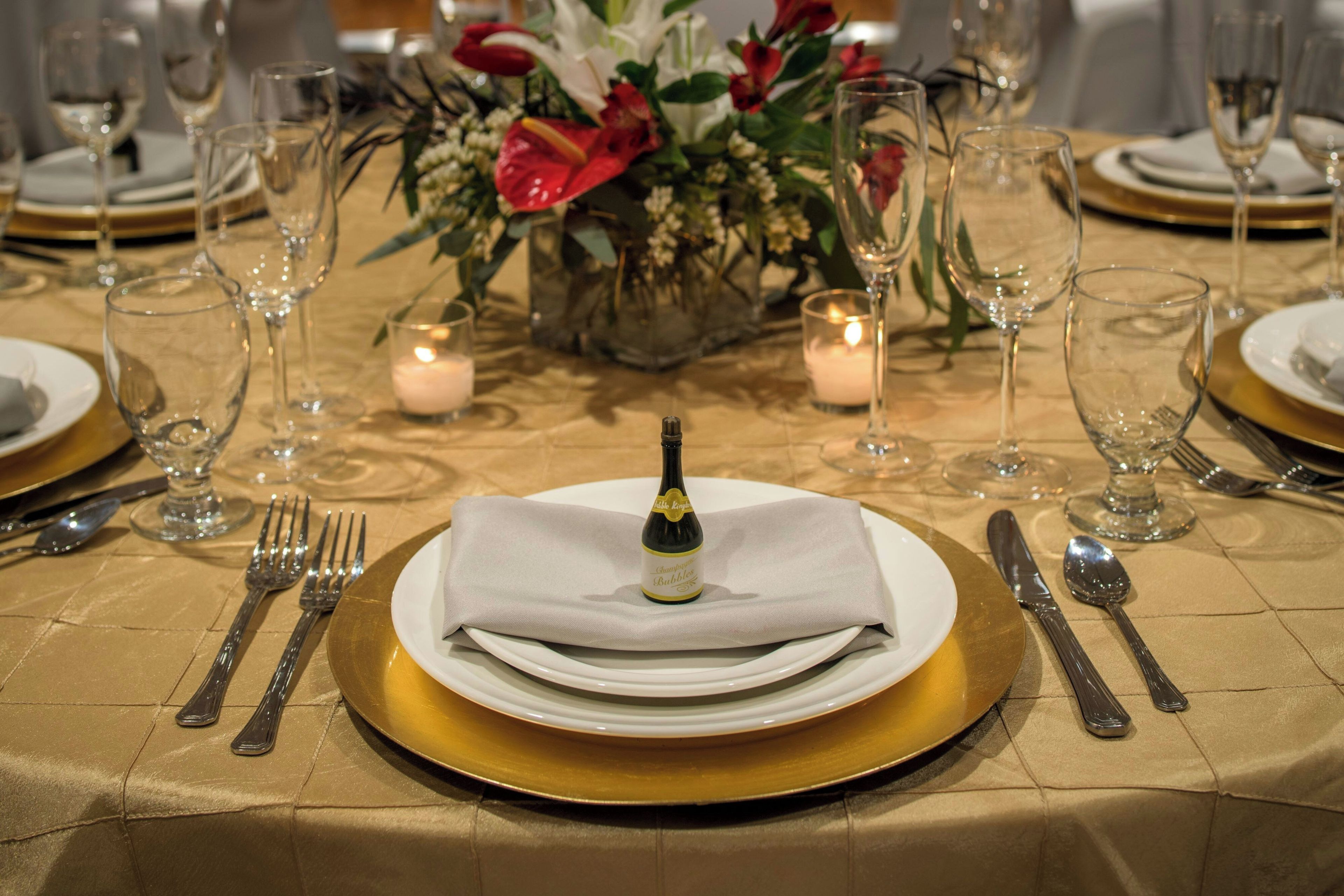A formal dining table set with gold and white tableware, a bottle of champagne, and a floral centerpiece.