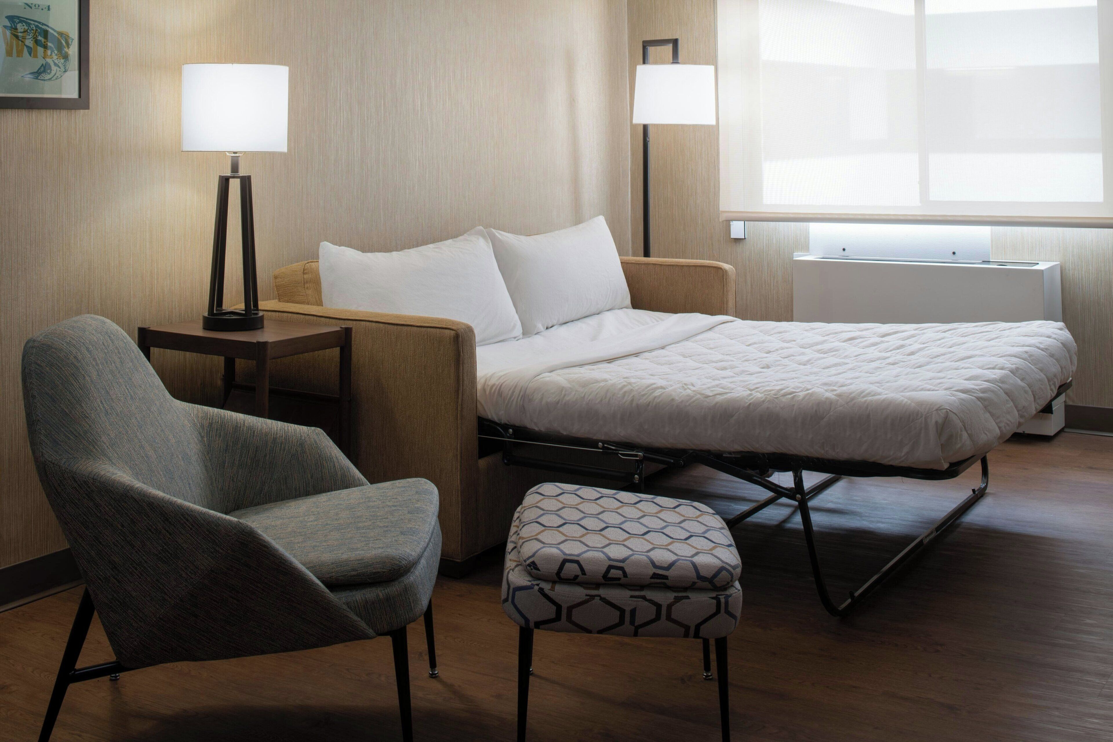 A hotel room with a bed, a chair, a small table, two lamps, and a window.