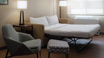 A hotel room with a bed, a chair, a small table, two lamps, and a window.
