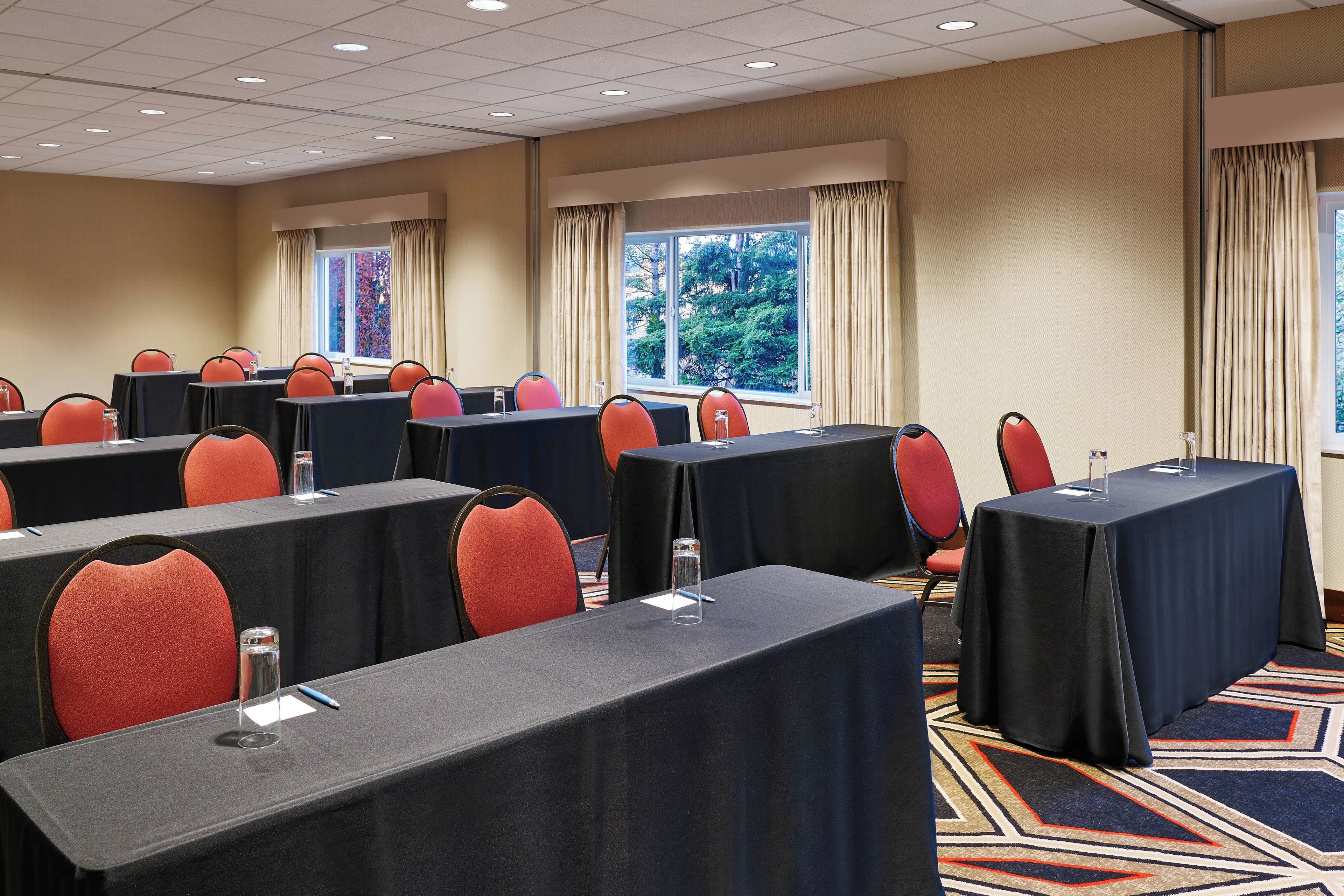 Un salón de conferencias con varias mesas y sillas, amplias ventanas con cortinas y piso alfombrado.