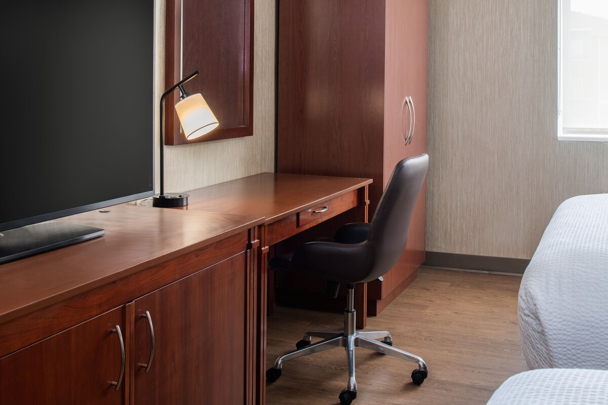 A hotel room with a wooden desk, a black office chair, a flat-screen TV, and a bed with white bedding.