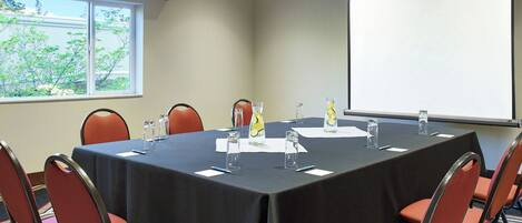 Un salón de conferencias con una mesa larga, sillas, una pantalla de proyector y una ventana con vistas a árboles.