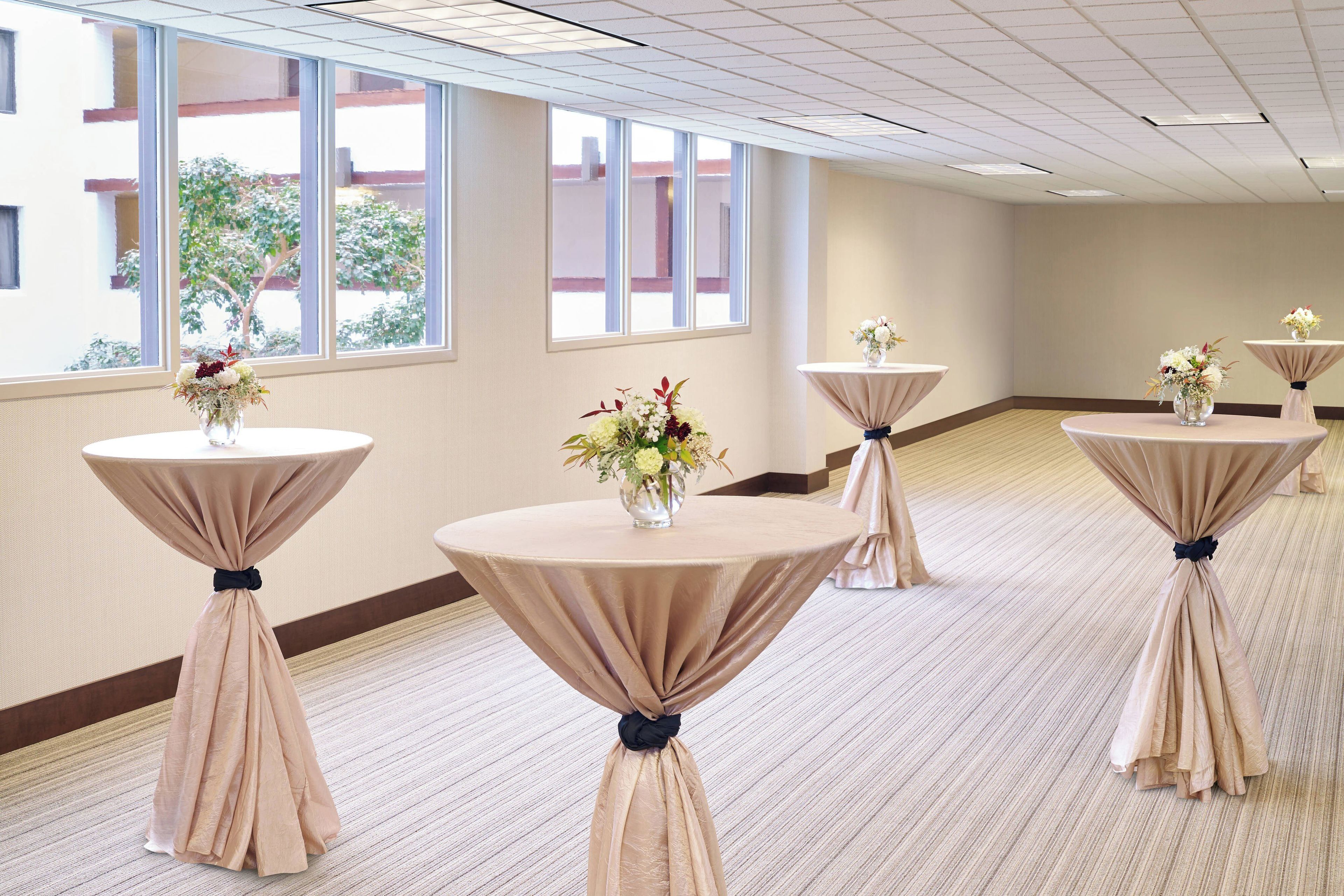 A hotel room with multiple round tables, each with a tablecloth and a vase of flowers.