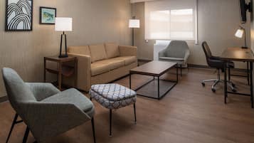 Ein modernes Hotelzimmer mit einer Couch, zwei Sesseln, einem Couchtisch und einem Schreibtisch mit Stuhl. Es gibt zwei Wandlampen und einen Flachbildfernseher.