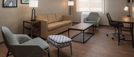 A modern hotel room with a sofa, two armchairs, a coffee table, and a desk with a chair. There are two wall-mounted lamps and a flat-screen TV.