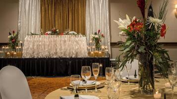 Un salón de banquetes con un escenario, mesas dispuestas para un evento formal y centros de mesa florales.