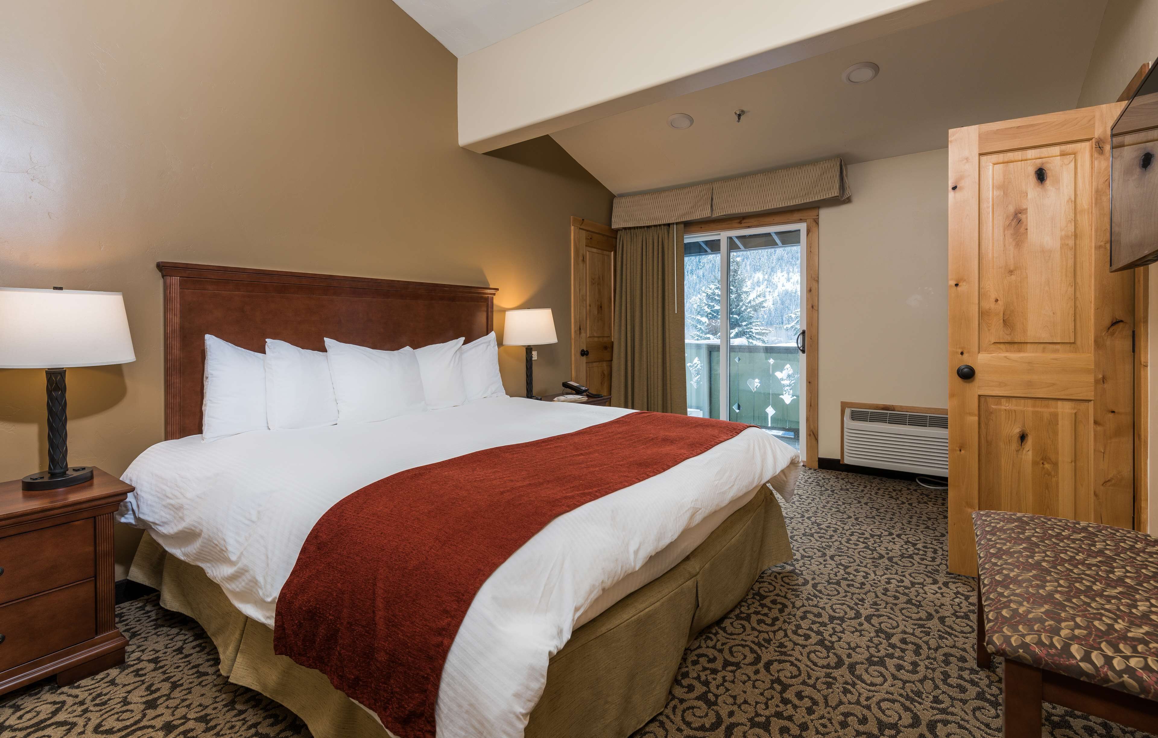 A hotel room with a large bed, two bedside lamps, a wooden headboard, a red throw blanket, a patterned carpet, a wooden chair, and a sliding glass door leading to a balcony.