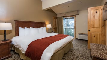 A hotel room with a large bed, two bedside lamps, a wooden headboard, a red throw blanket, a patterned carpet, a wooden chair, and a sliding glass door leading to a balcony.