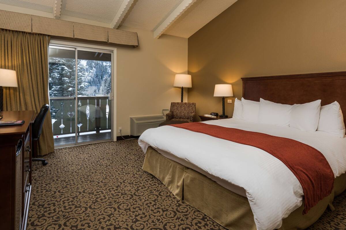 A hotel room with a large bed, two bedside lamps, a chair, a desk, and a view of a snowy landscape through a sliding door.
