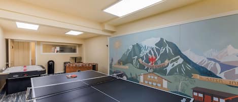 A room with a ping pong table, a foosball table, and a mural depicting mountains and a lodge.