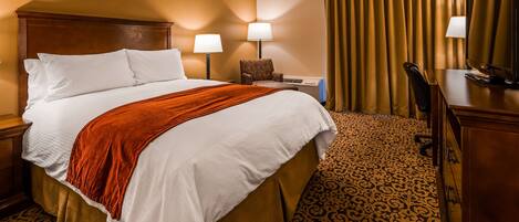 A hotel room with a large bed, two bedside lamps, a chair, a desk, a television, and a patterned carpet.