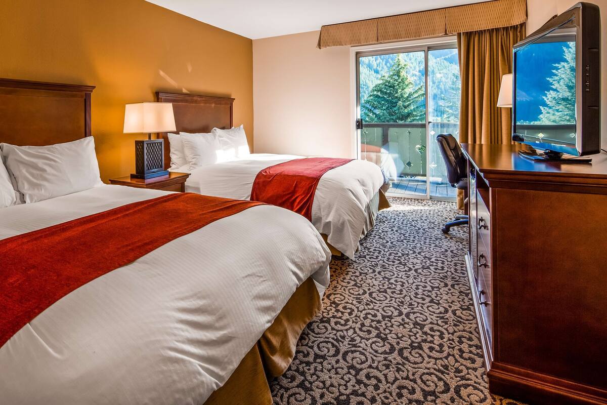 A hotel room with two beds, a desk, a television, and a view of trees through the window.