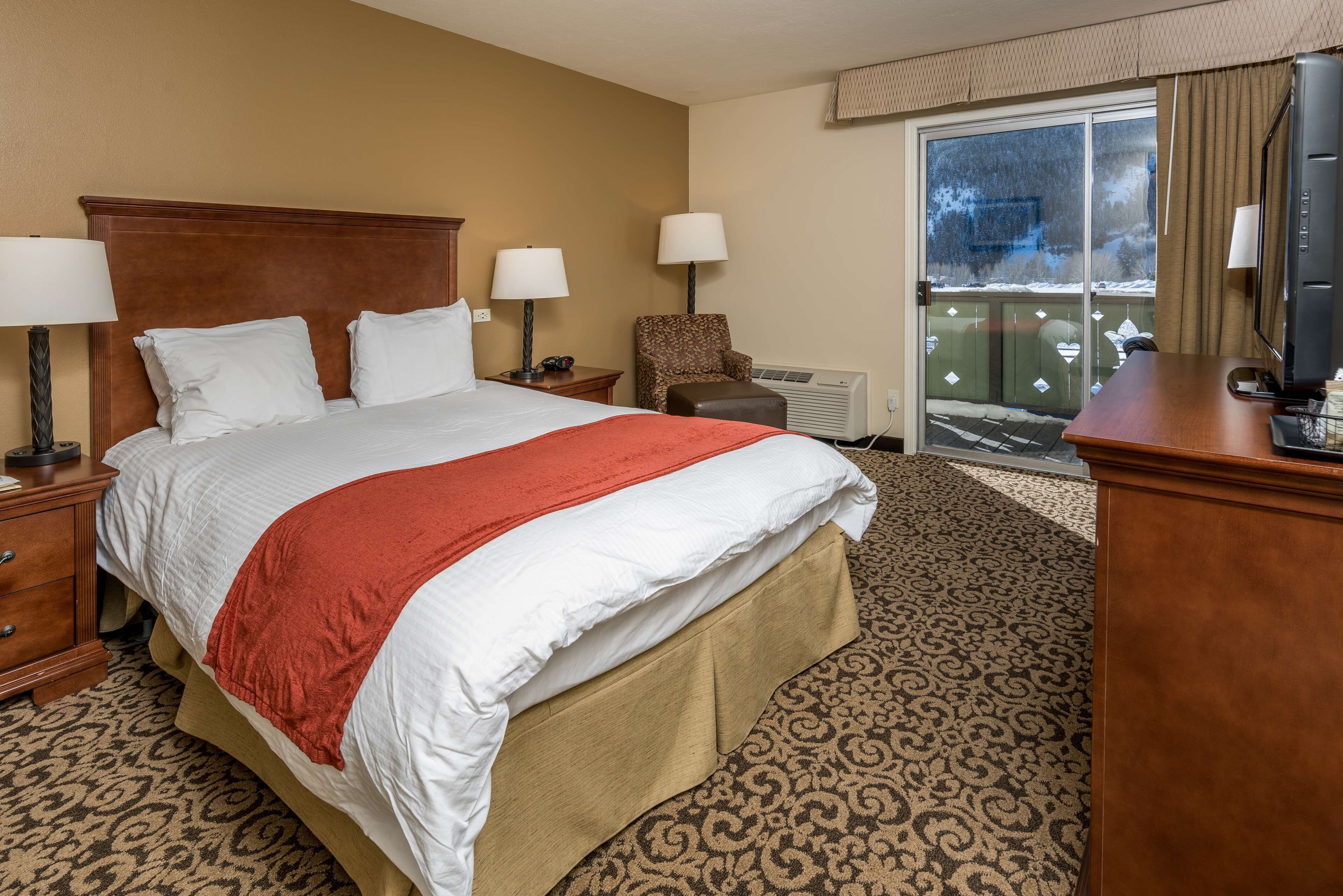 A hotel room with a large bed, two bedside tables with lamps, a chair, a desk with a TV, and a sliding glass door to the outside.