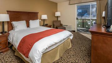 A hotel room with a large bed, two bedside tables with lamps, a chair, a desk with a TV, and a sliding glass door to the outside.