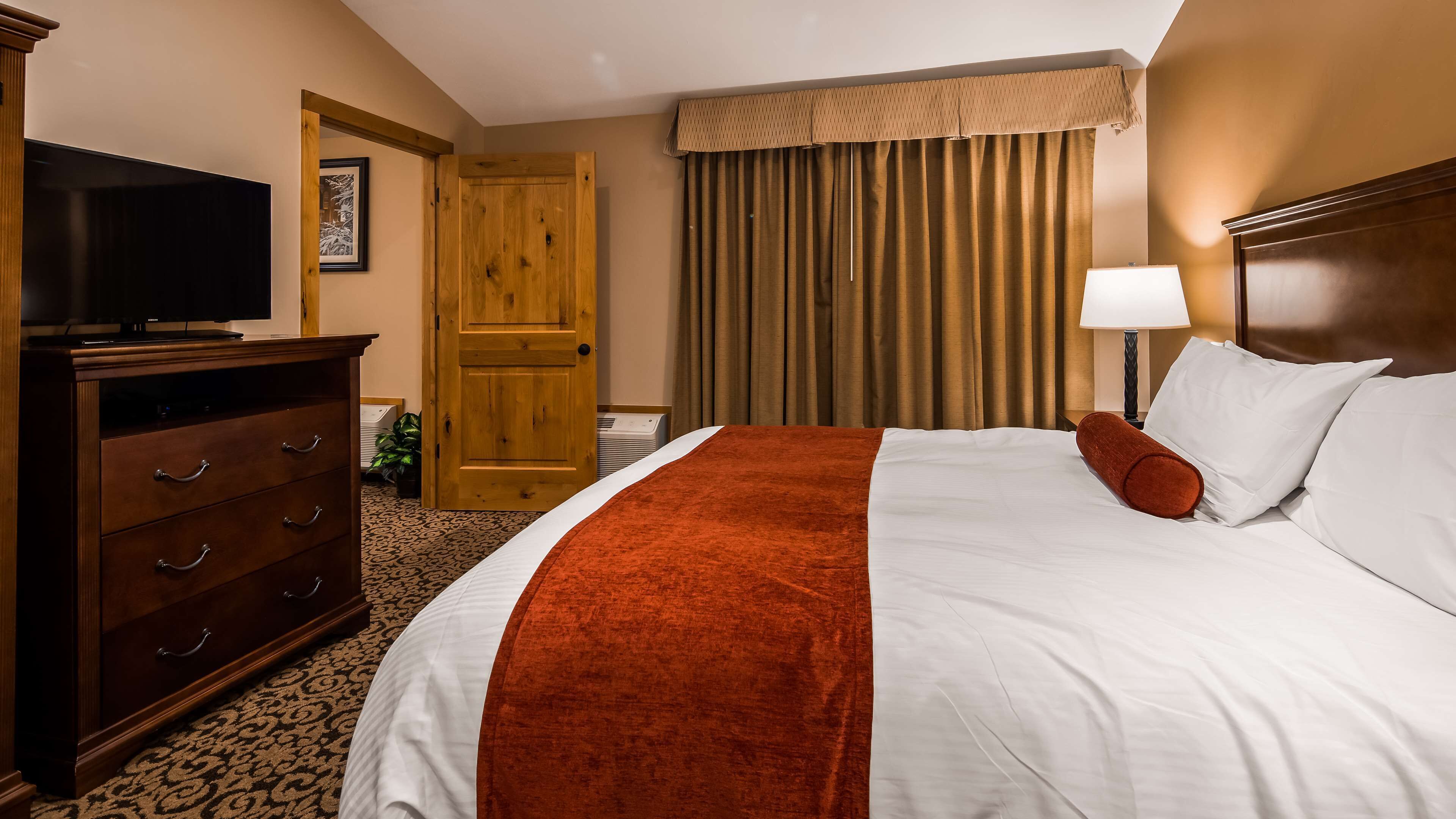 Habitación de hotel con cama, televisión, armario de madera, puerta y ventana con cortinas.