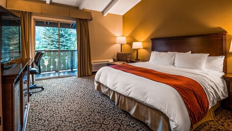 A hotel room with a large bed, a desk with a chair, a balcony with a view of trees, and a lamp on the nightstand.