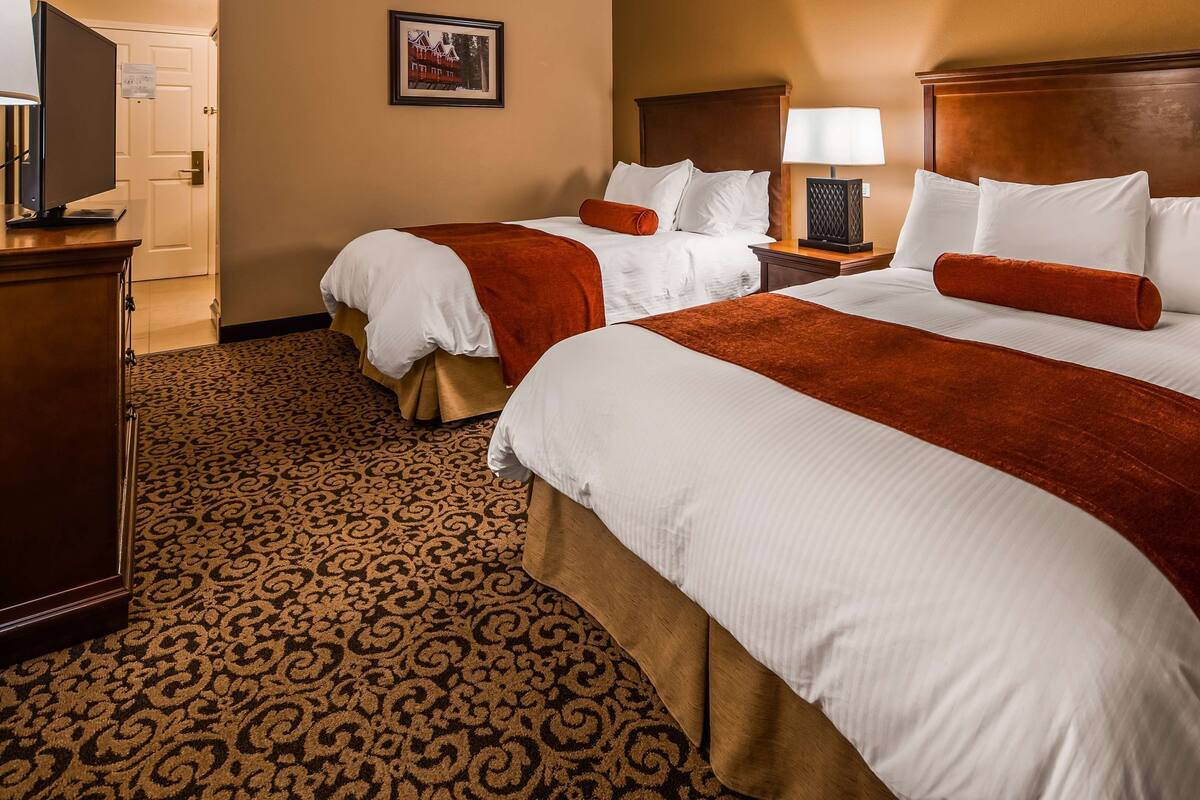 A hotel room with two beds, each with white linens and red throw pillows, a wooden headboard, a nightstand with a lamp, and a patterned carpet.