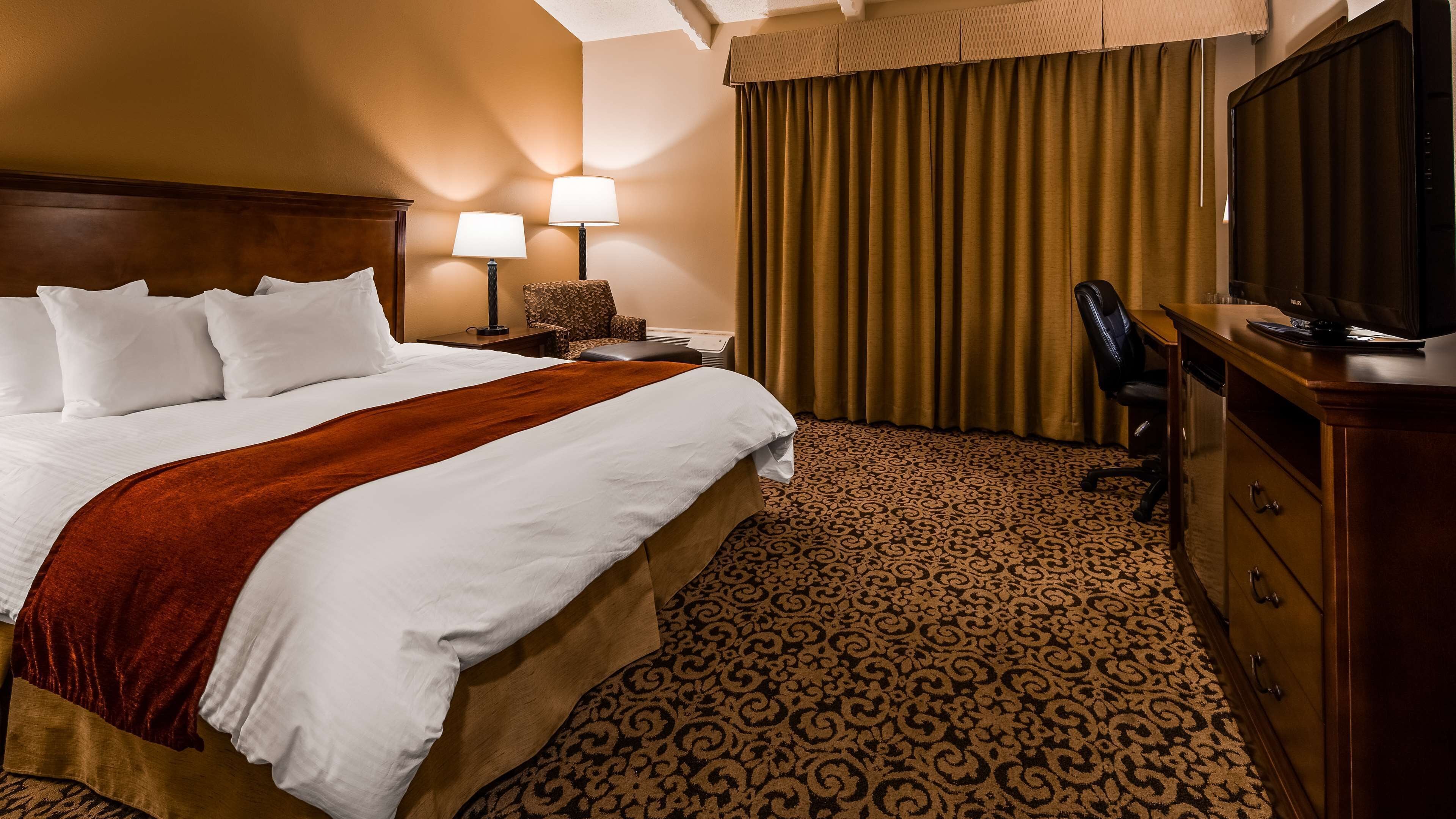 A hotel room with a large bed, two bedside lamps, a desk with a chair, a television, and a patterned carpet.