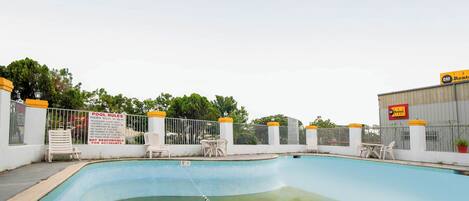 A swimming pool with a pool rule sign, white chairs, and a fence.