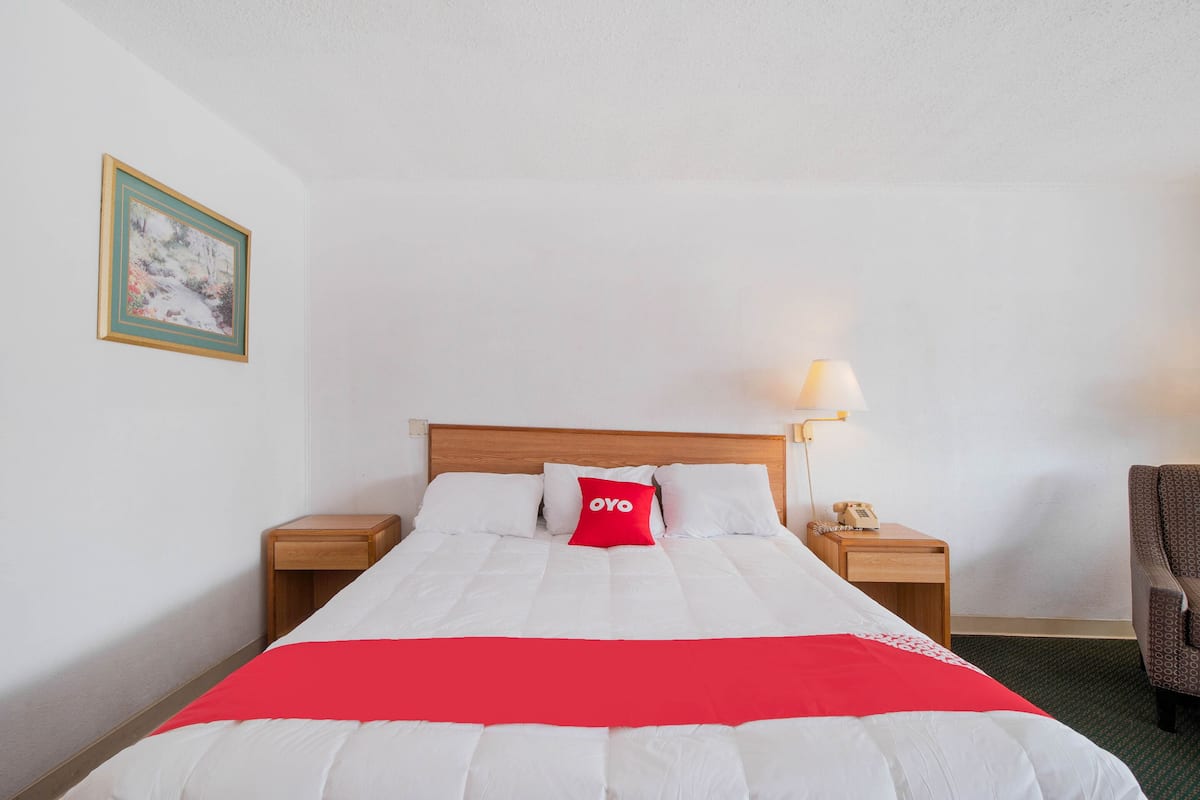 Habitación de hotel con cama, mesita de noche con lámpara, una silla e imagen enmarcada en la pared.