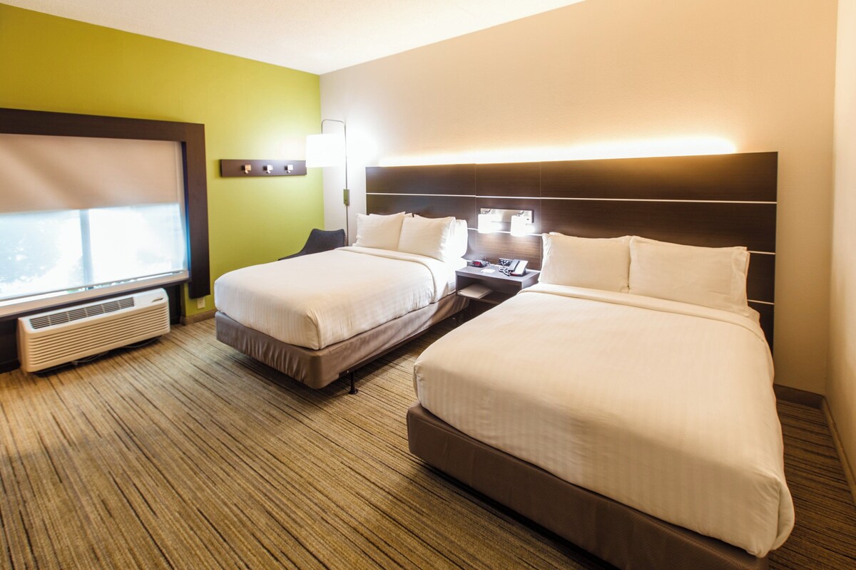 A hotel room with two beds, a window, a heater, and a nightstand with a lamp.