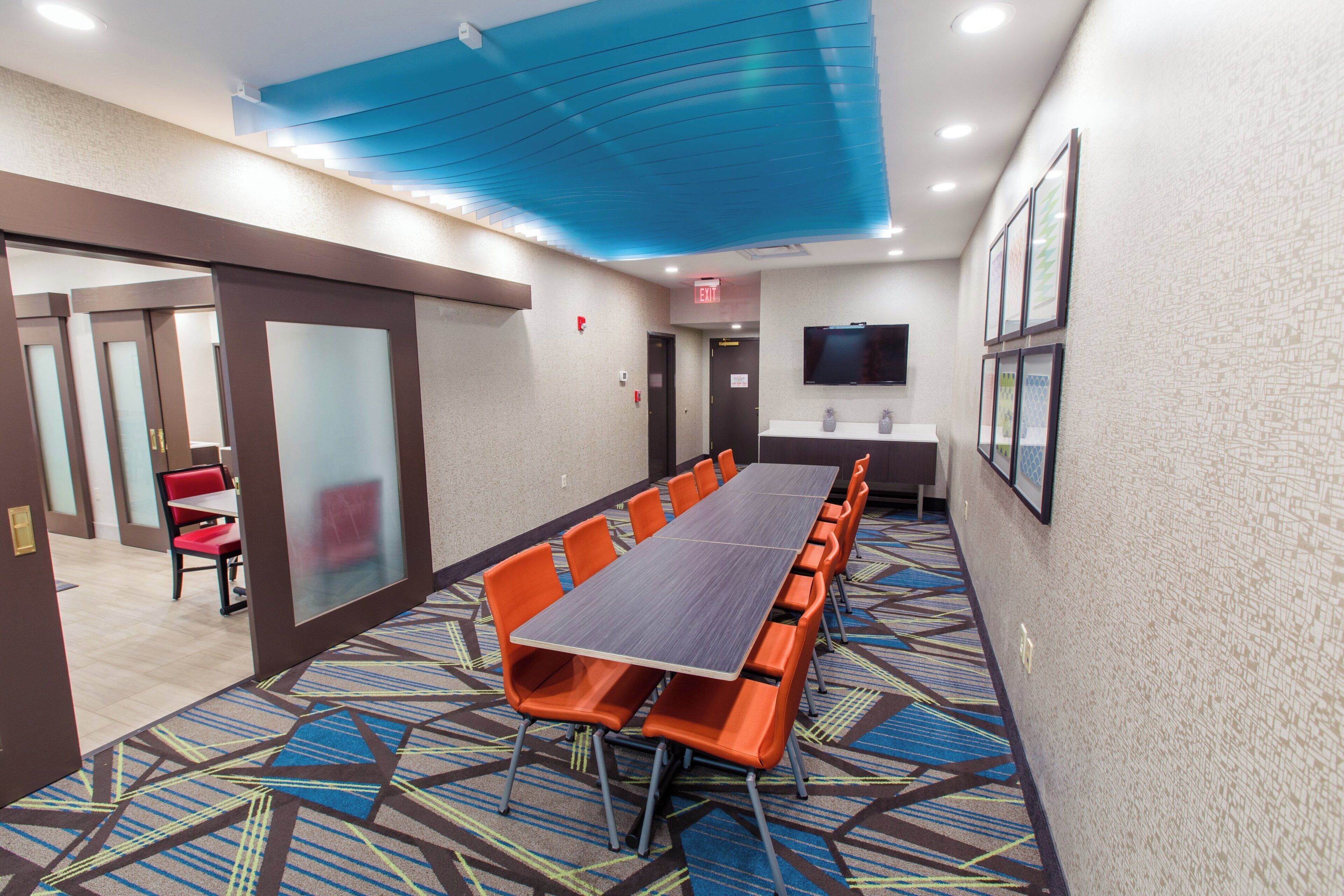 Un salón de conferencias con una mesa larga, sillas color naranja, un televisor de pantalla plana y una alfombra estampada.