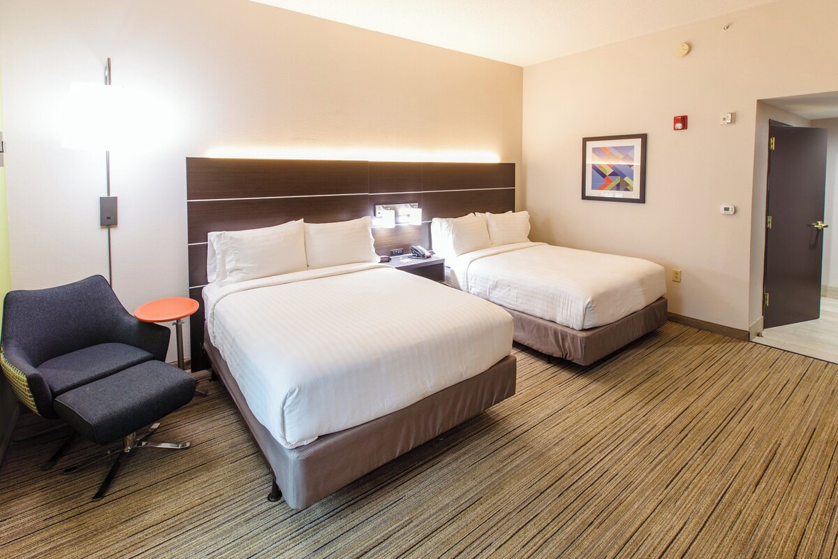 A hotel room with two beds, a chair, a small table, and a wall-mounted lamp.
