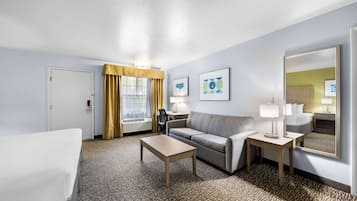 A hotel room with a bed, a sofa, a small table, a desk, a chair, a lamp, a mirror, and artwork on the wall.