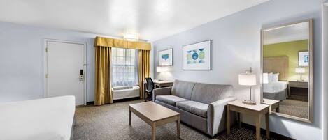 A hotel room with a bed, a sofa, a small table, a desk, a chair, a lamp, a mirror, and artwork on the wall.