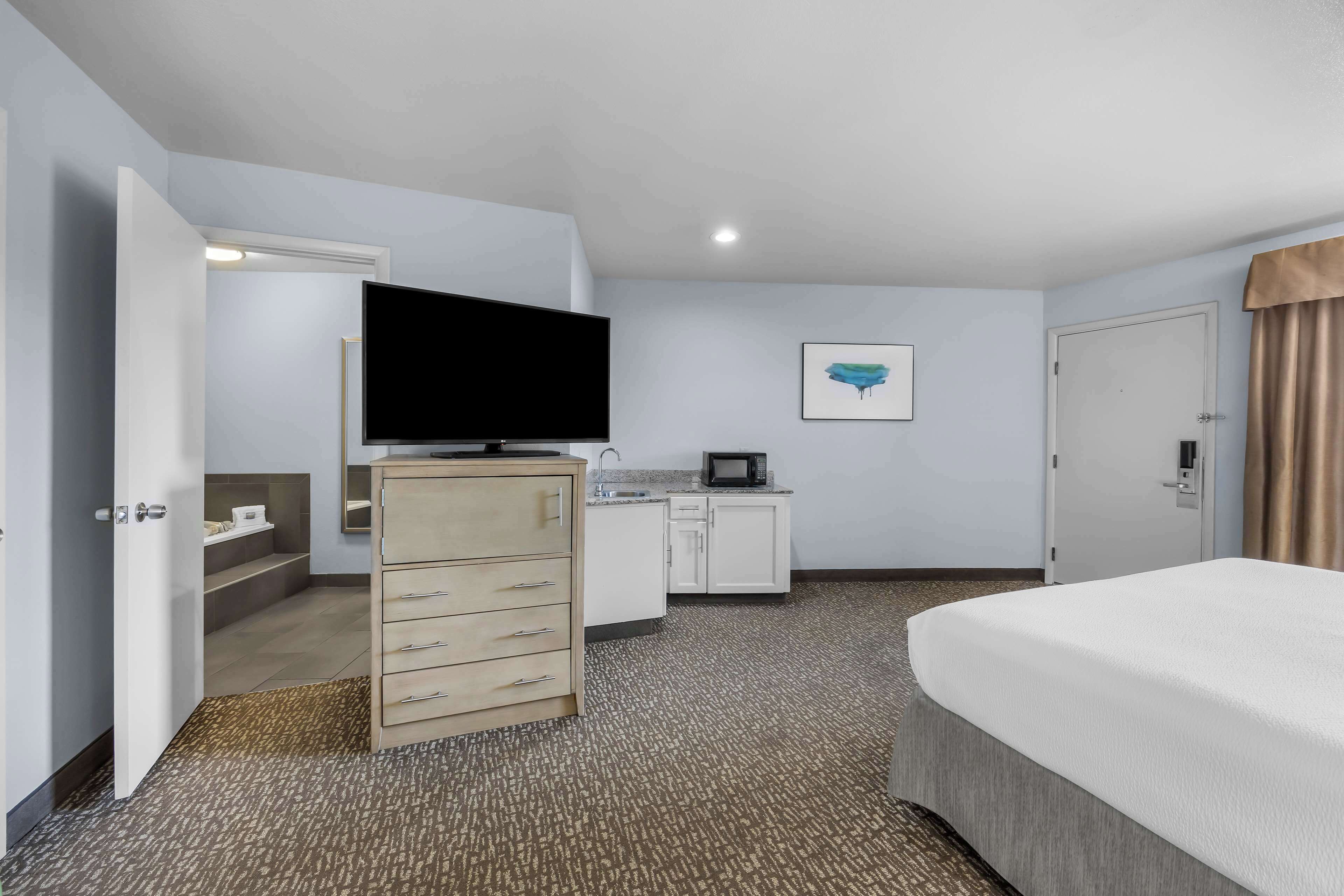 Habitación de hotel con cama, televisión, escritorio con microondas, cómoda y baño con lavamanos e inodoro.