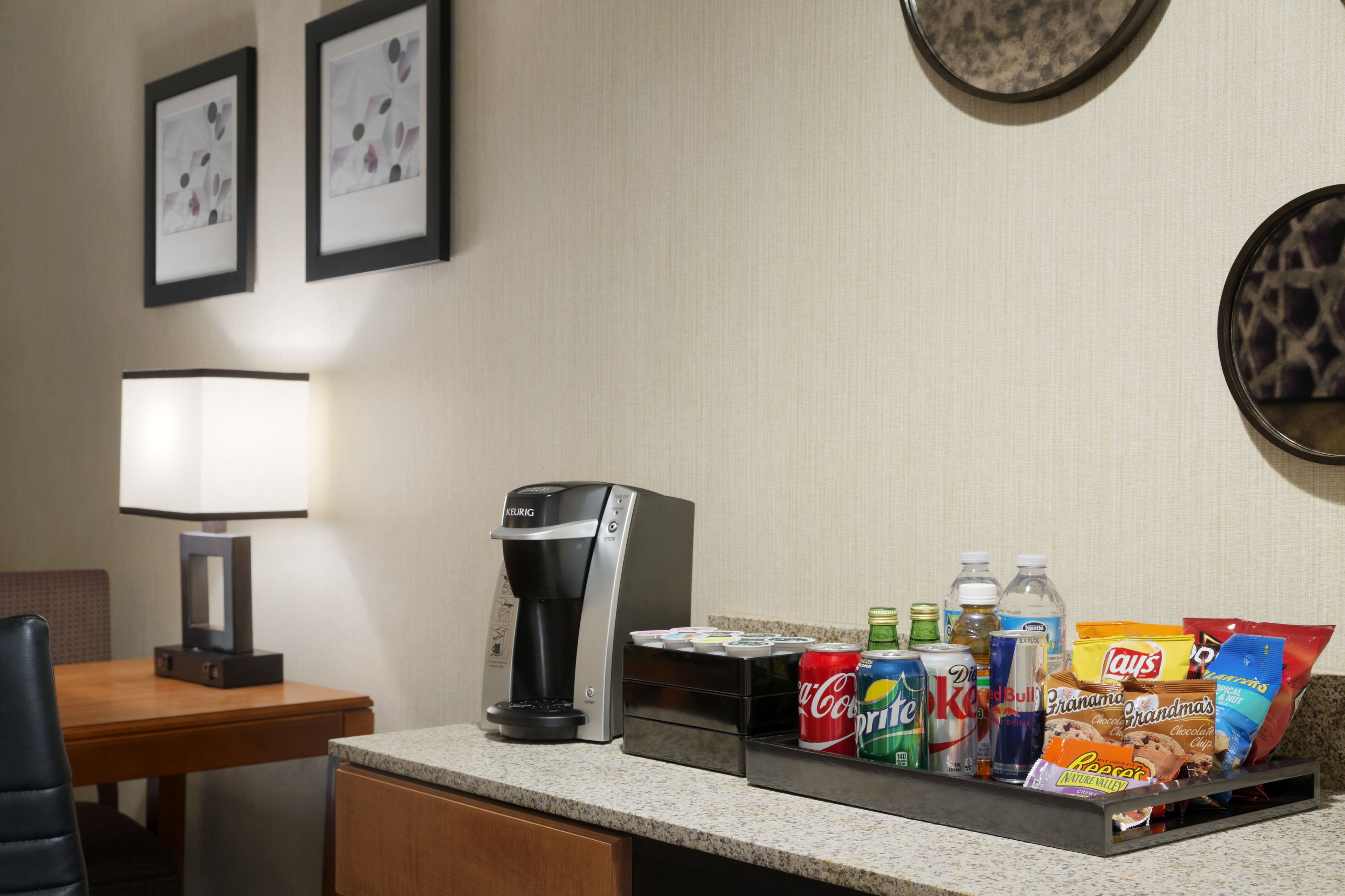 Une chambre d’hôtel équipée d’une machine à café, d’un plateau avec des collations et des boissons, d’une lampe et de tableaux encadrés au mur.