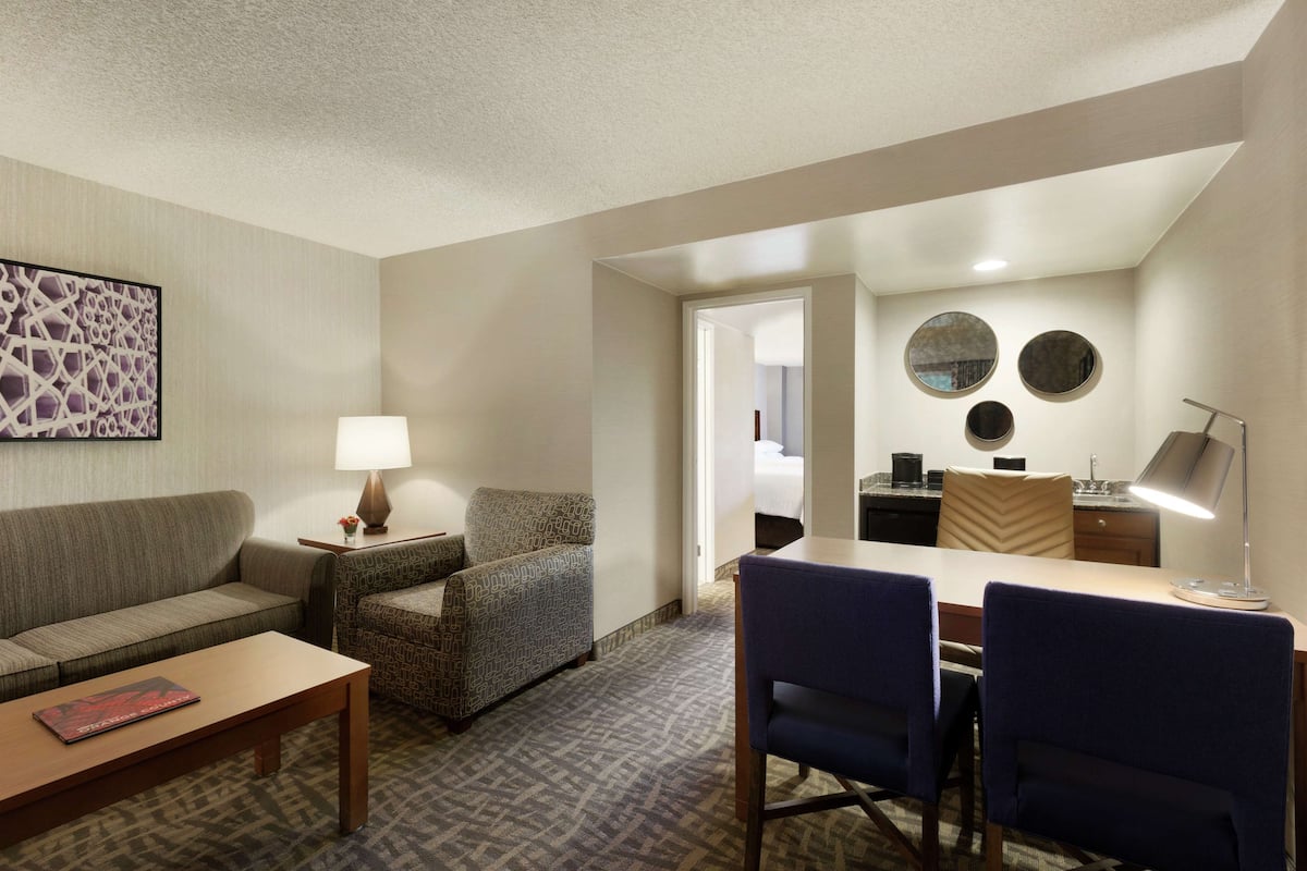 A hotel room with a sofa, armchair, dining table, and chairs. There is a lamp on the table and a painting on the wall.