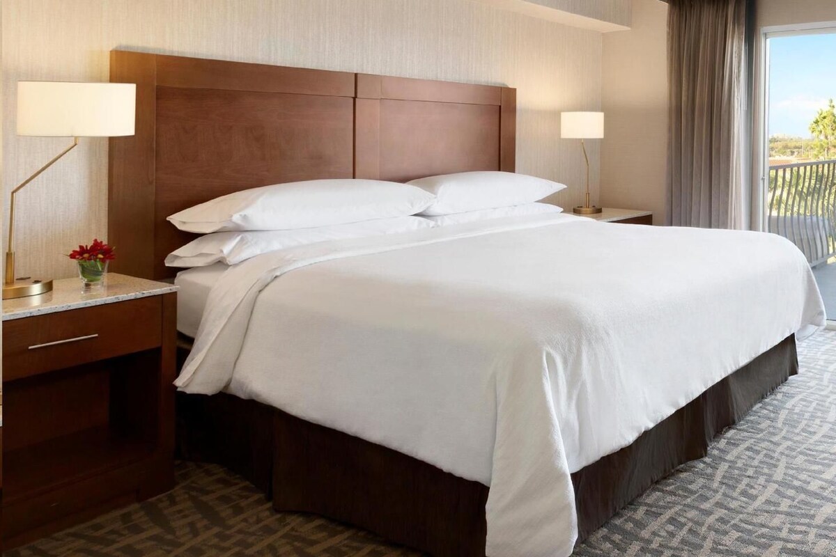 Habitación de hotel con una cama grande, dos mesitas de noche, cortinas y vistas al exterior.