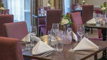 A dining area with tables set for two, featuring red upholstered chairs, clear glassware, and white napkins.