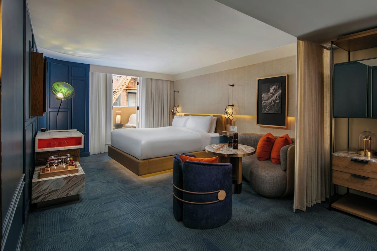 A hotel room with a bed, a seating area with a table, a TV, and a minibar.