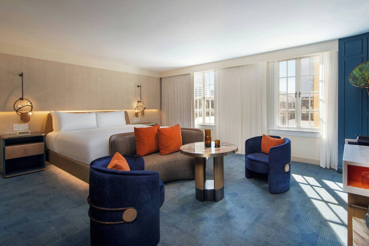 A modern hotel room with a bed, a sofa, two armchairs, a small table, and a nightstand.