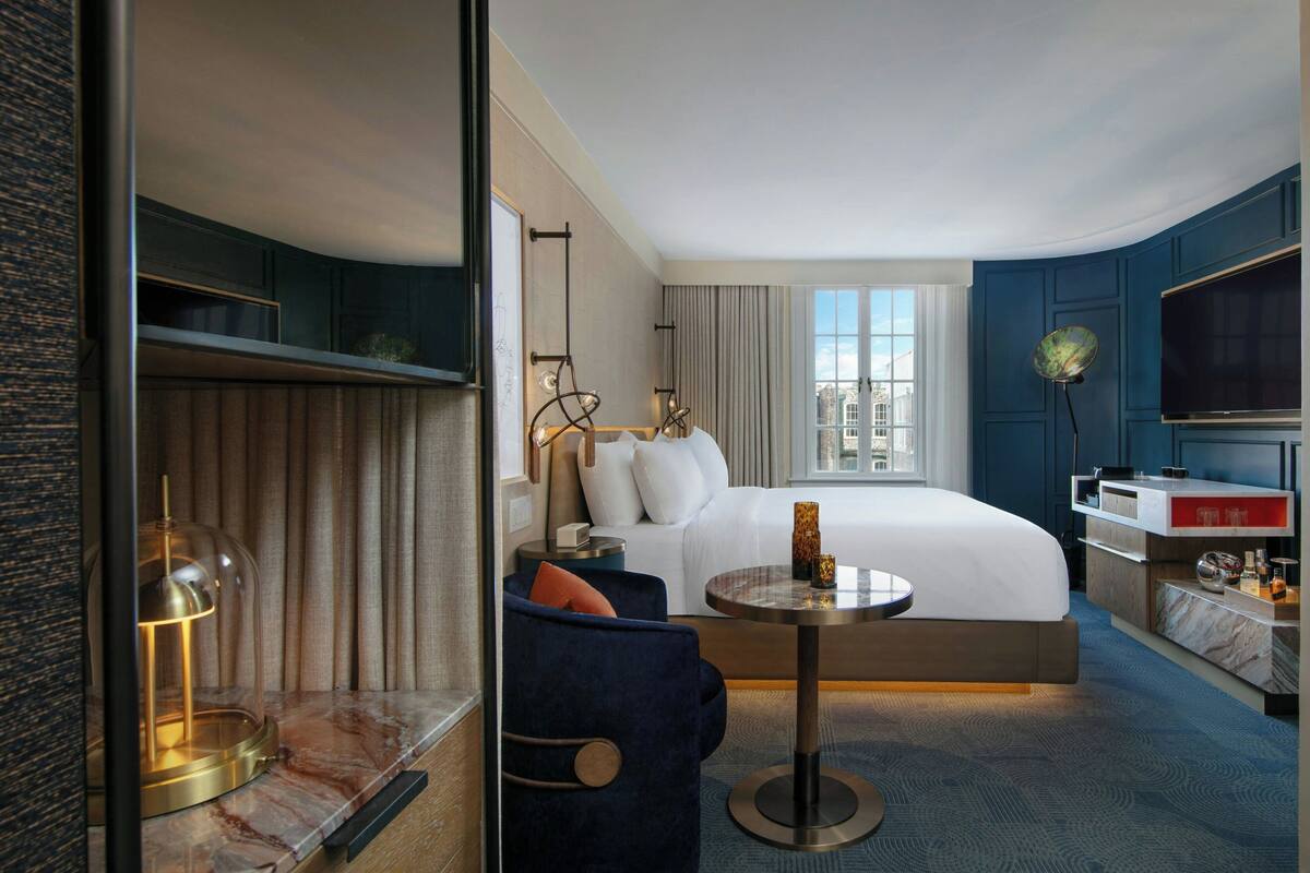 A modern hotel room with a large bed, a round table, a blue armchair, a TV, and a view of the city.