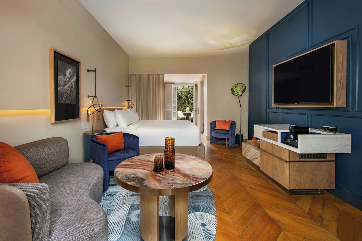 A modern hotel room with a bed, a seating area with a round table, a TV, and a kitchenette.