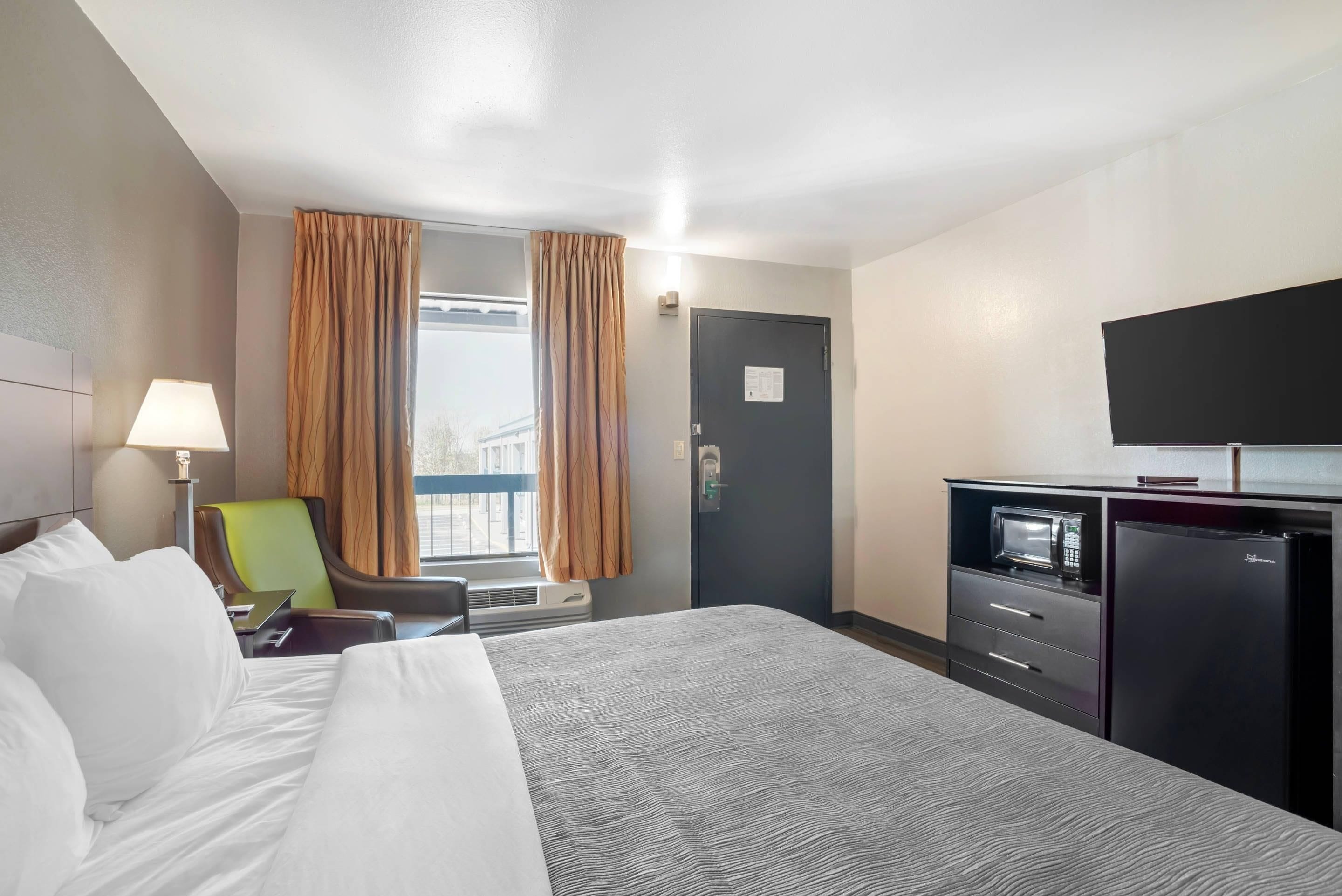 Una camera d'albergo con un letto, una scrivania con forno a microonde, una sedia, una finestra con tende e una televisione a parete.