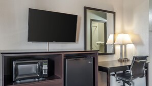 A hotel room with a flat-screen TV, a minibar, a desk with a chair, and a lamp.