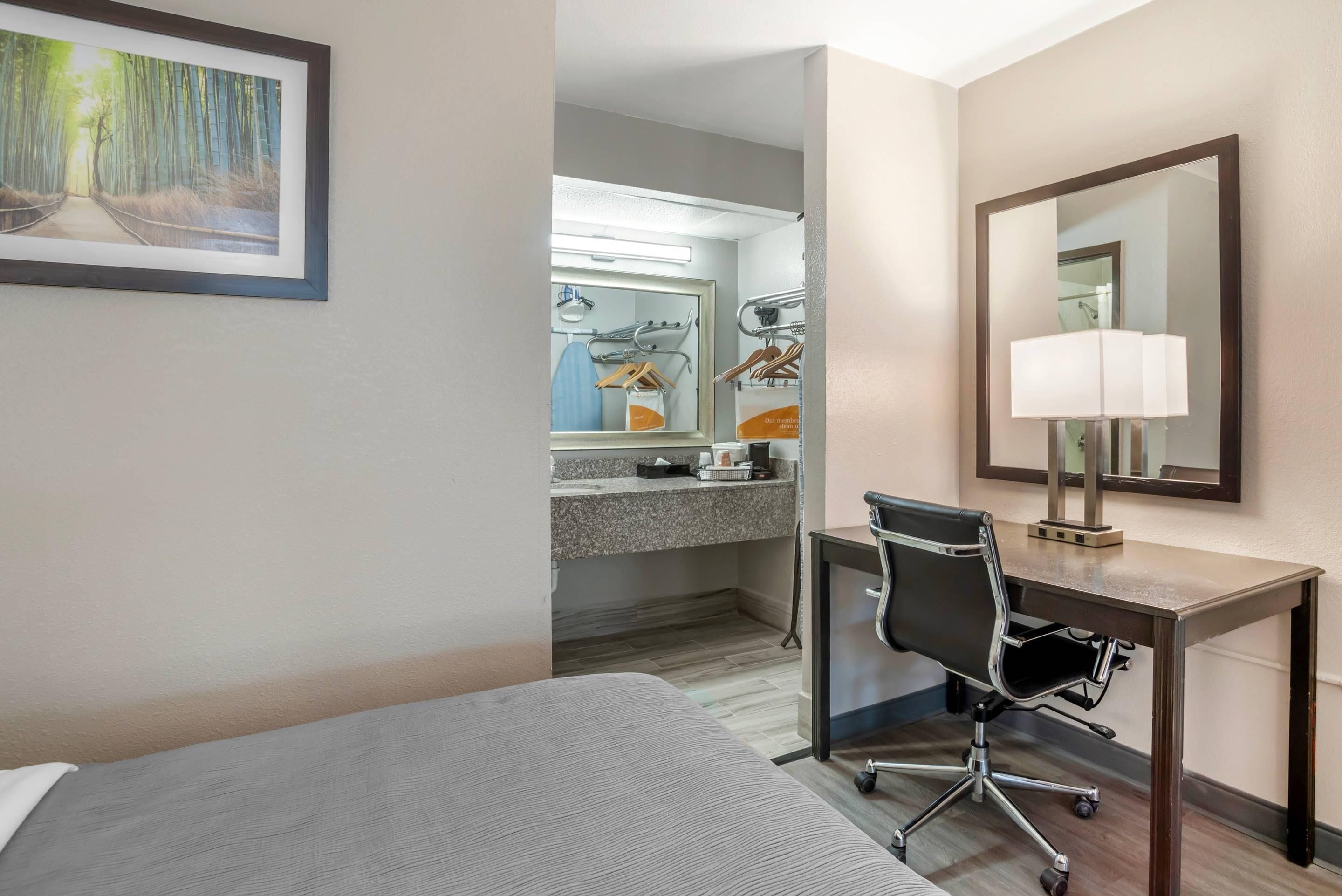 Habitación de hotel con cama, escritorio, silla, espejo y baño visible a través de una puerta abierta.