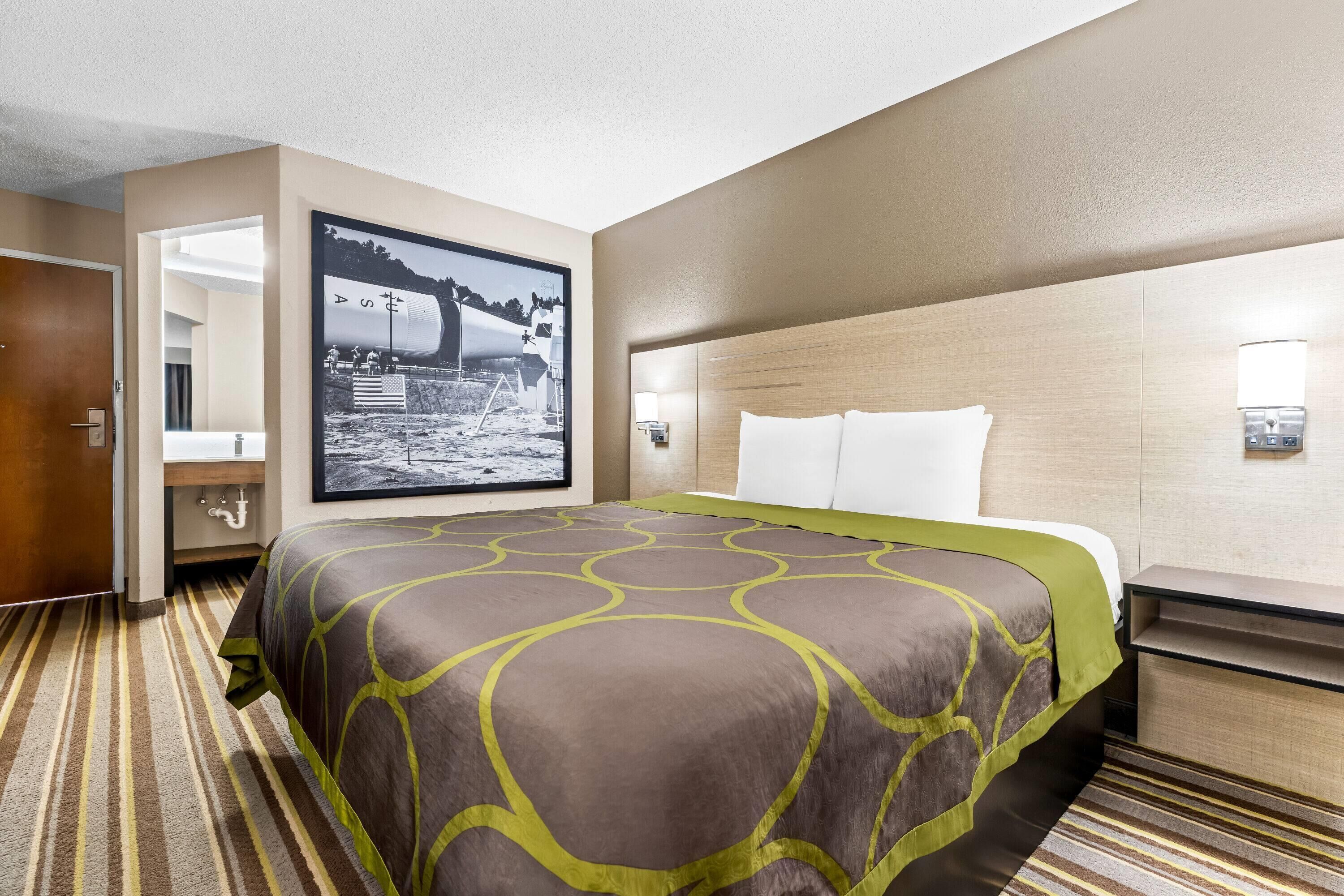 A hotel room with a large bed, a headboard, a bedside table, a nightstand, a wall-mounted lamp, and a framed picture on the wall.
