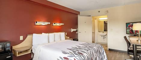 A hotel room with a bed, bedside table, lamp, desk, chair, and a bathroom visible through an open door.